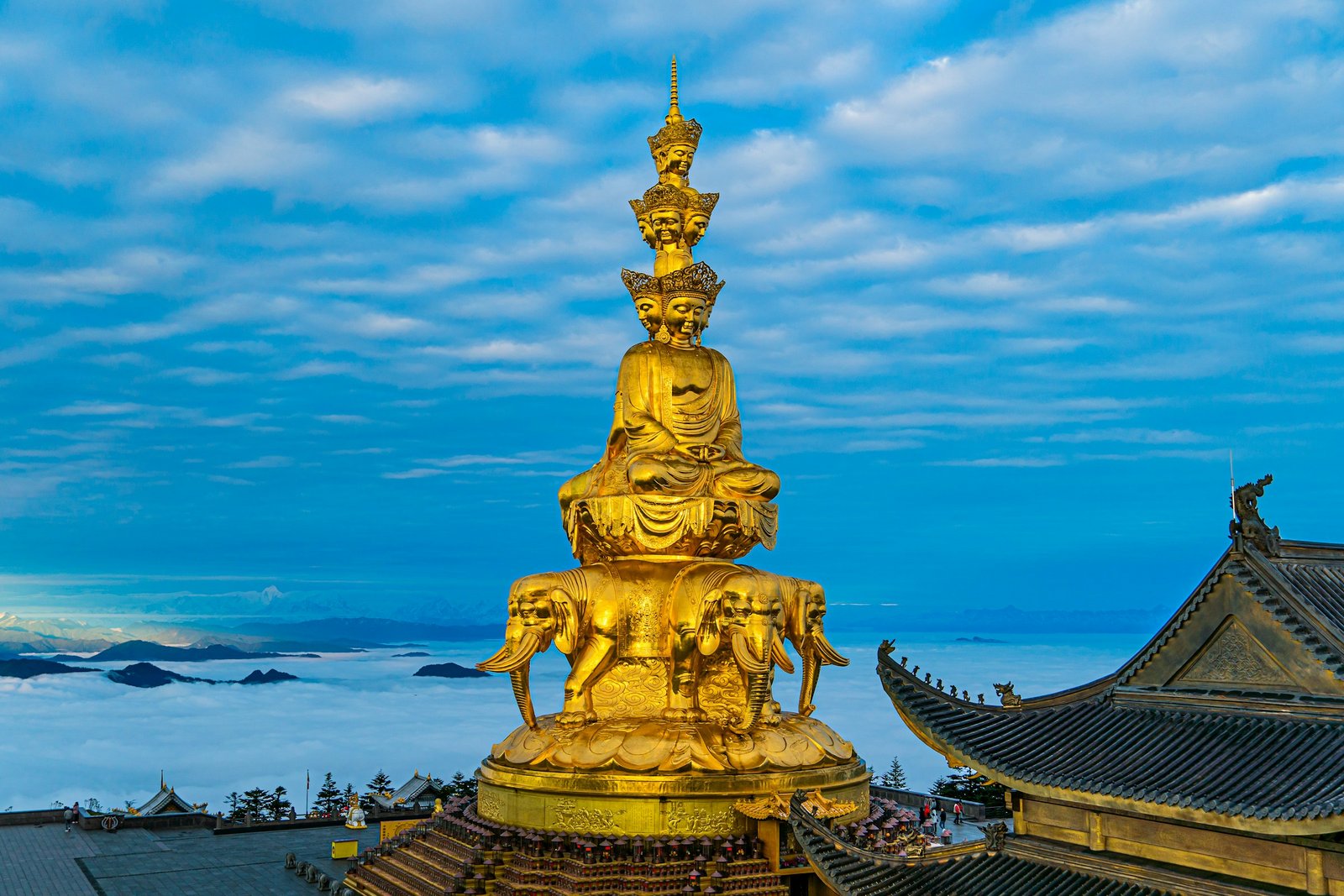 The complete golden Samantabhadra statue above a sea of clouds on Mount Emei, with temple roofs and small visitors around its base.